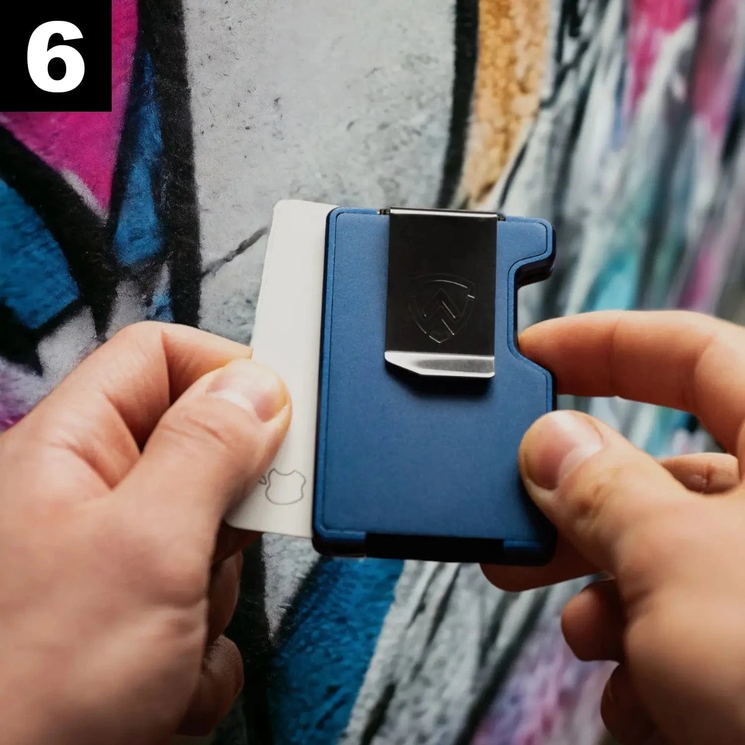 Person holding a blue minimalist wallet with a money clip against a colorful graffiti wall.